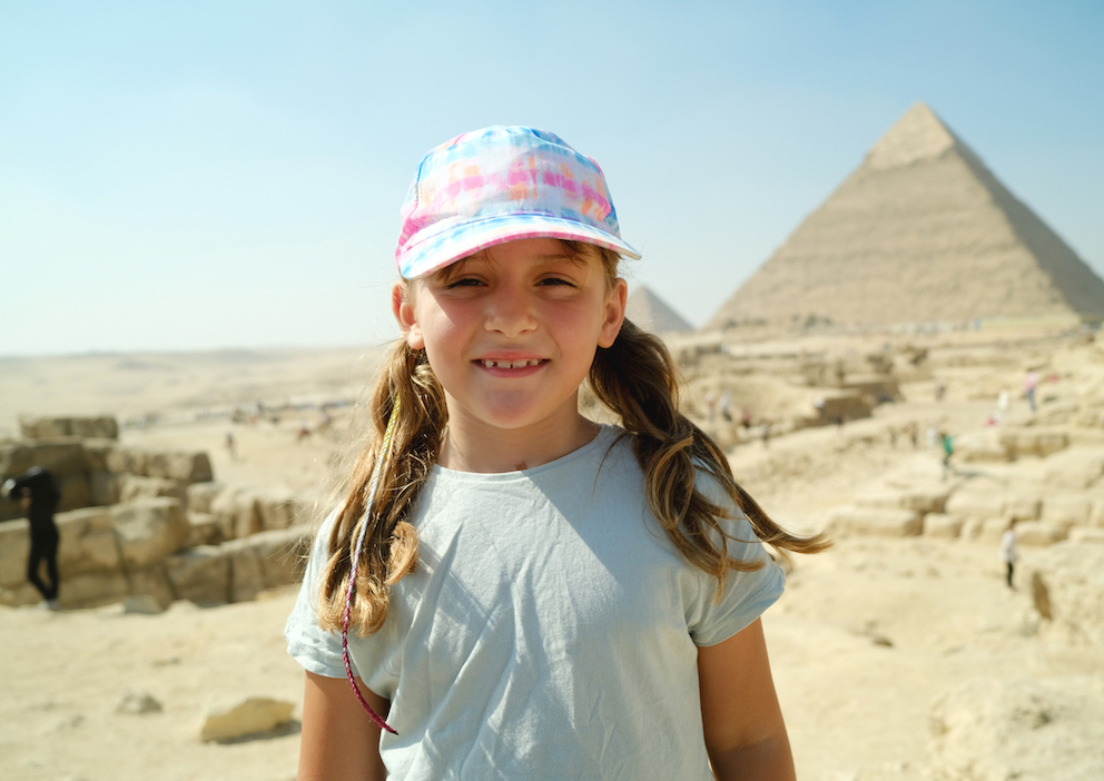 Vor einer Pyramide steht ein Mädchen mit zwei Zöpfen und einer Kappe.