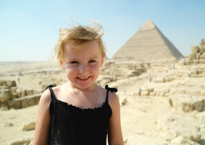 Vor einer Pyramide steht ein blondes Mädchen. Es lächelt in die Kamera.